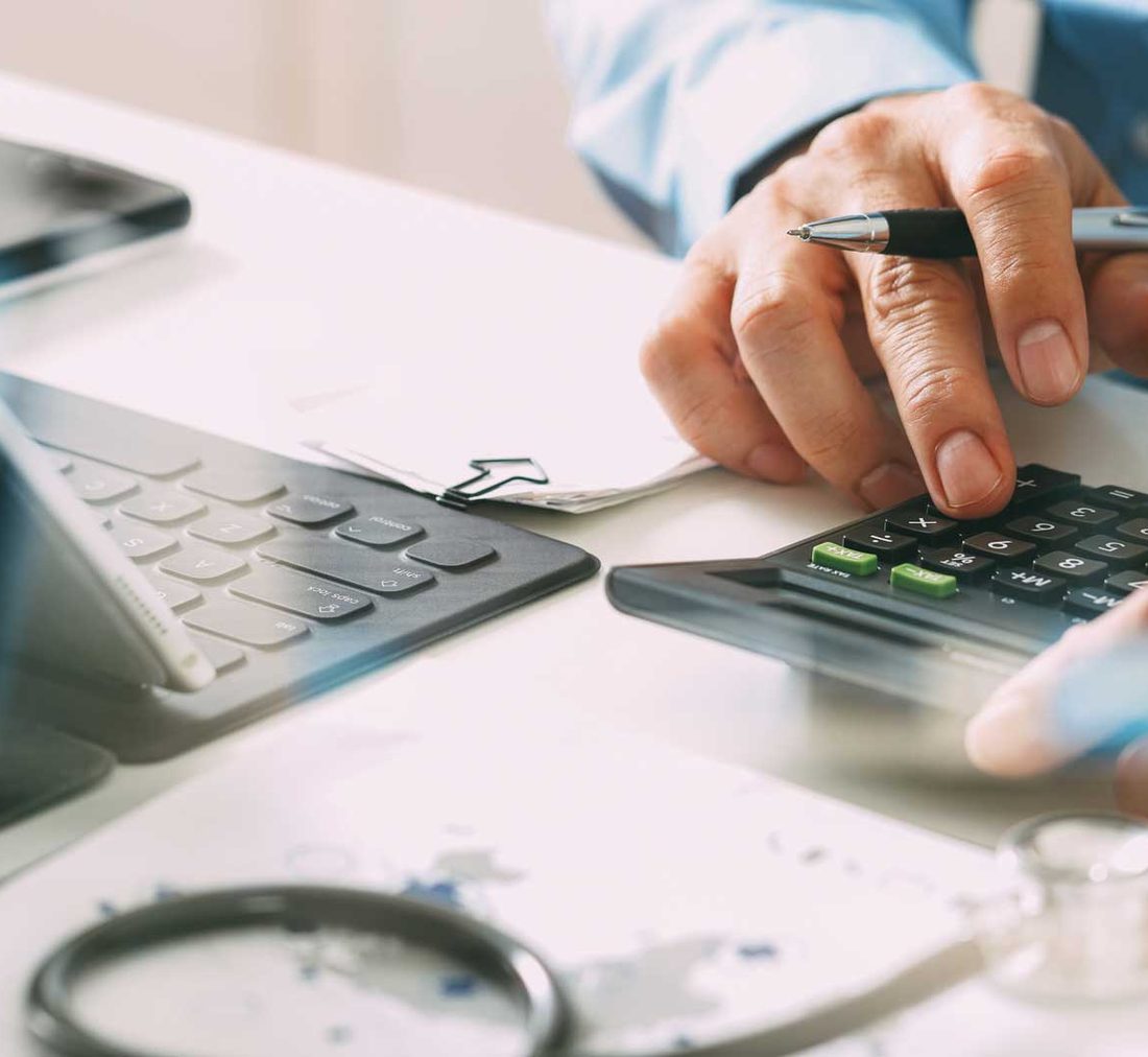 A finance professional working through patient account figures on a calculator, a stethoscope on the desk beside them